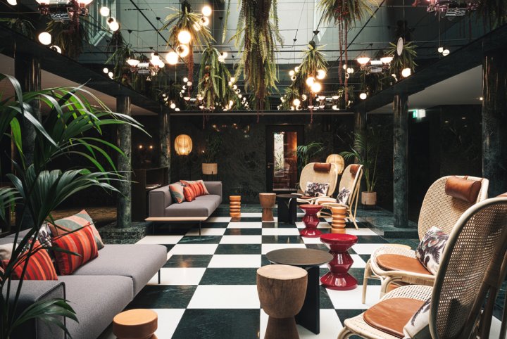 Hotellobby mit Schachbrettboden und grünen Marmorsäulen, möbliert mit grauen Sofas und Stühlen mit natürlichem Geflecht, Pflanzen sowie runde Lampen hängen von der Decke.