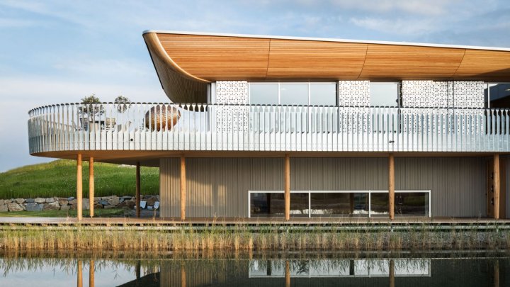 Ein schiffartiges Haus mit Holzdach und einem Balkon rund um das erste Geschoss mit Holzpfosten vor einem See
