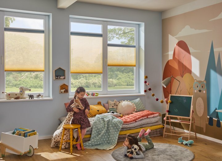 Zwei kleine Mädchen spielen in einem bunten Kinderzimmer mit Wabenplissees als Sonnenschutz an den Fenstern