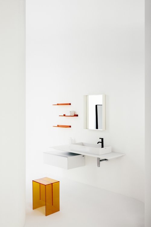 Weisses Waschbecken mit rechteckigem, weissem Spiegel darüber, daneben drei leuchtend orange Wandregale und ein oranger Hocker im Vordergrund.