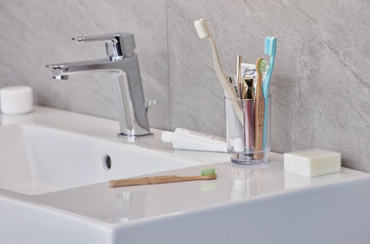 Zahnputzglas mit diversen Zahnbürsten und Utensilien steht auf einem weissen Waschbecken mit silbernem Wasserhahn.