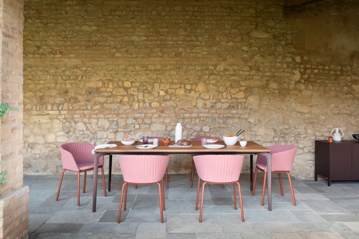 Gedeckter Tisch aus dunklem Holz mit rosa Stühlen in einer Outdoornische mit Natursteinwänden.