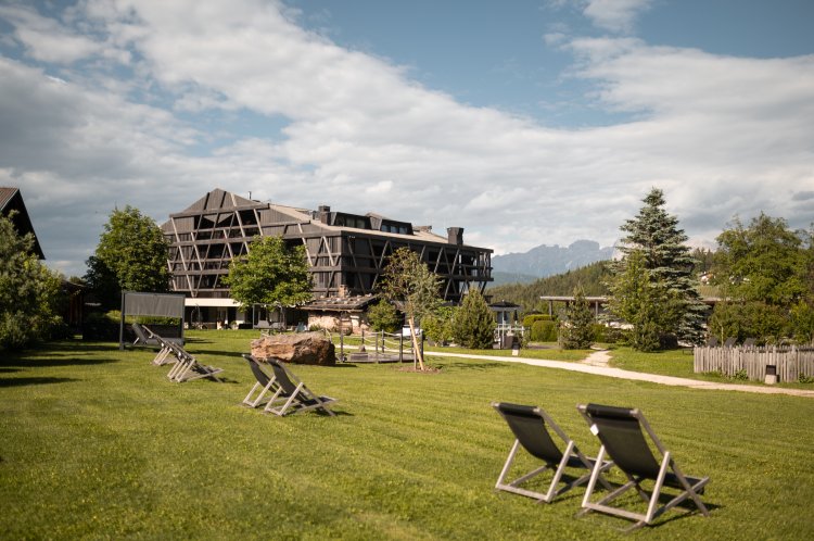 Hotel im modernen Chalet-Stil auf grüner Wiese mit Liegestühlen, im Hintergrund Berge und blauer Himmel