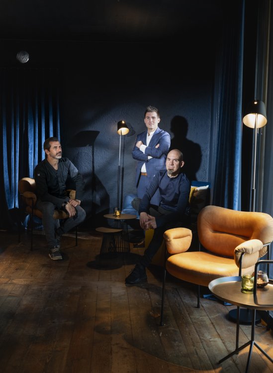 Andreas Caminada Sitzend links mit Giuseppe lo Vasco stehend in der Mitte und Timo Fritsche sitzend rechts vor dunkelblauem Vorhant und einem hellbrauen Lederstuhl in der Ecke im Restaurant Oz.