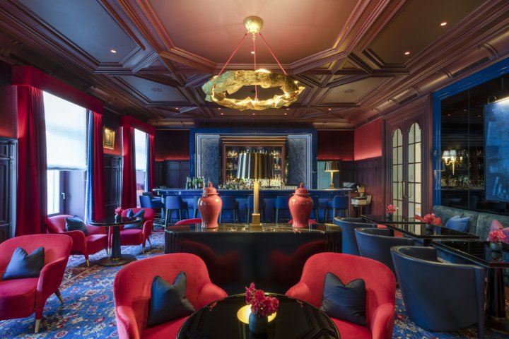 Gemütliche, edle Hotelbar mit goldenem Kronleuchter, stühlen in einem satten Rot, einem dunkelblauen Teppich und einer historischen Holzdecke.