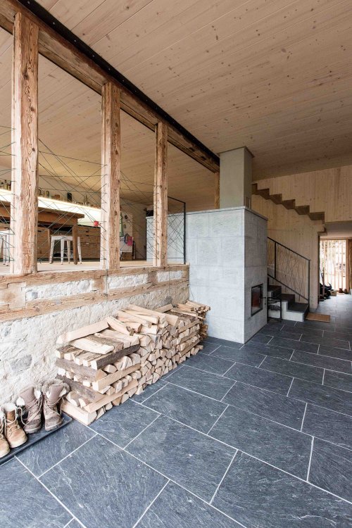 Eingangsbereich mit grauen Steinplatten offen zum leicht erhöhten Wohnzimmer, das mit alten Holzbalken abgetrennt ist.