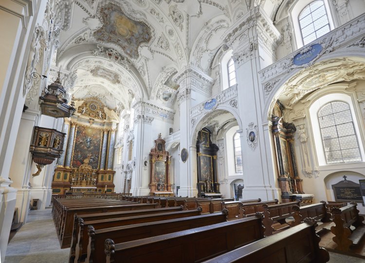 Innenraum der barocken Jesuitenkirche in Solothurn mit Kirchenbänken aus Holz im Vordergrund und weissen Stuckaturen im hohen Kirchenraum im Hintergrund.