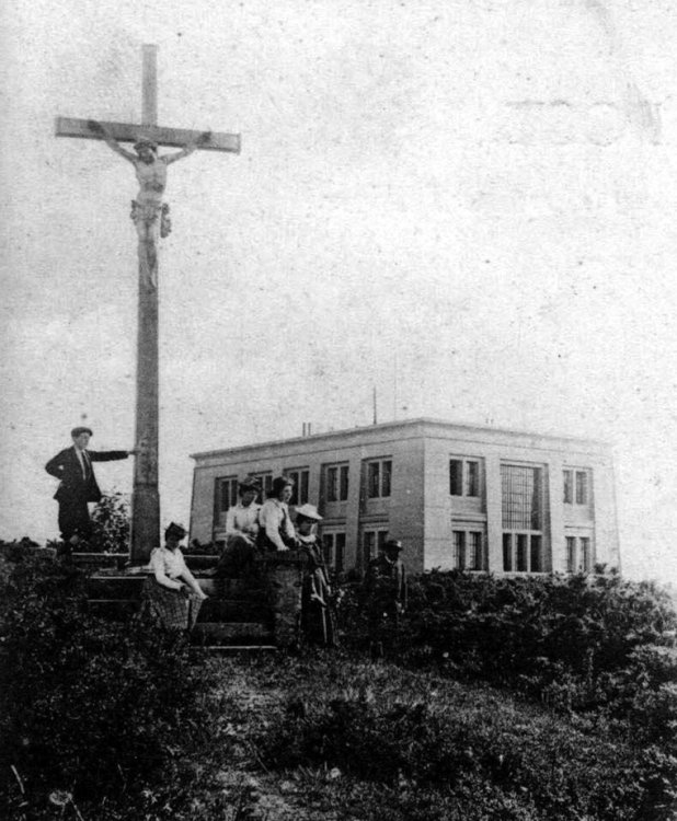 Schwarz-weiss Fotografie eines Hauses mit Flachdach, davor sitzen einige Menschen gruppiert um ein Kreuz mit Jesus.