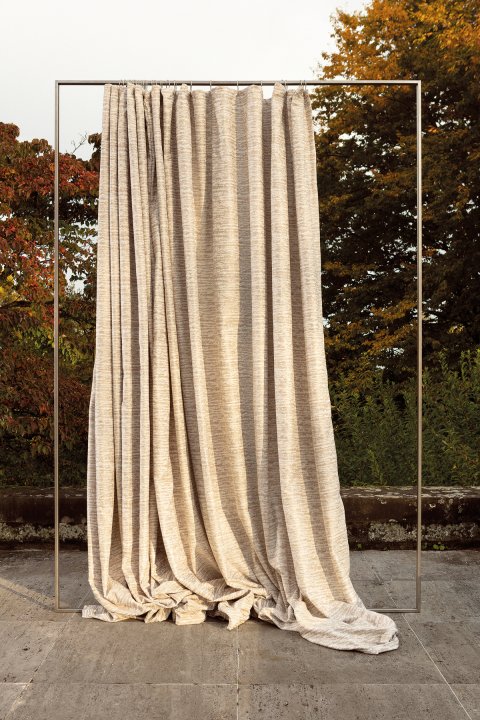 Beiger Vorhang in Baumrinde-Optik hängt an einer freistehenden, goldenen Vorhangstange im Freien vor herbstlichen Bäumen