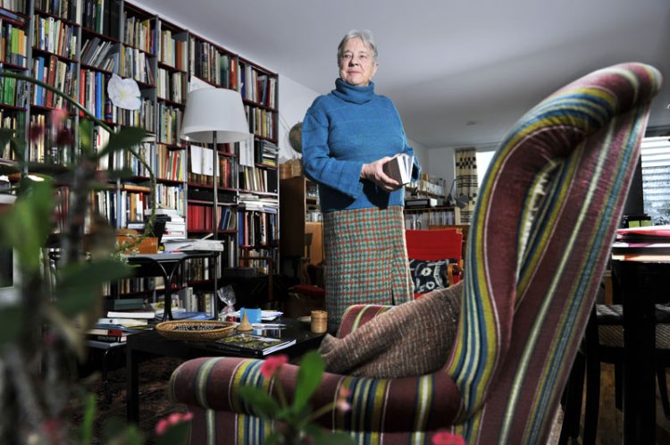 Verena Huber steht in einem blauen Pullover in ihrem Zuhause, im Vordergrund ein bunt gestreifter Sessel, hinter ihr ein volles Bücherregal