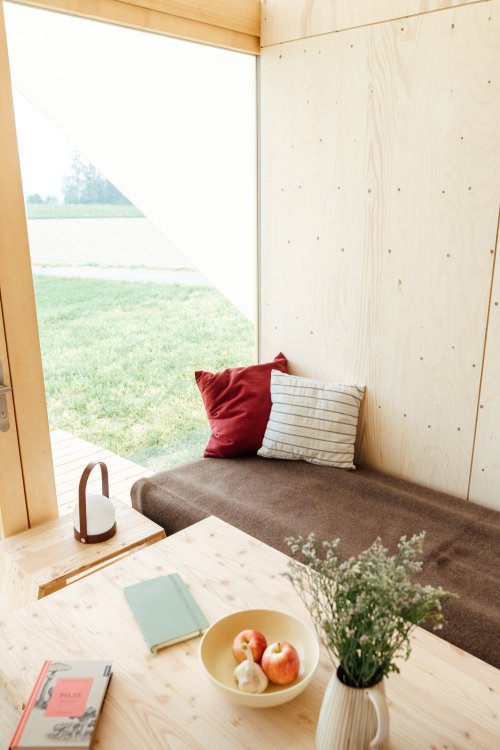 Braunes Sofa mit zwei Kissen steht am Fenster eines Holzhauses, davor ein Tisch mit Früchten, Notizbuch und Blumenstrauss.