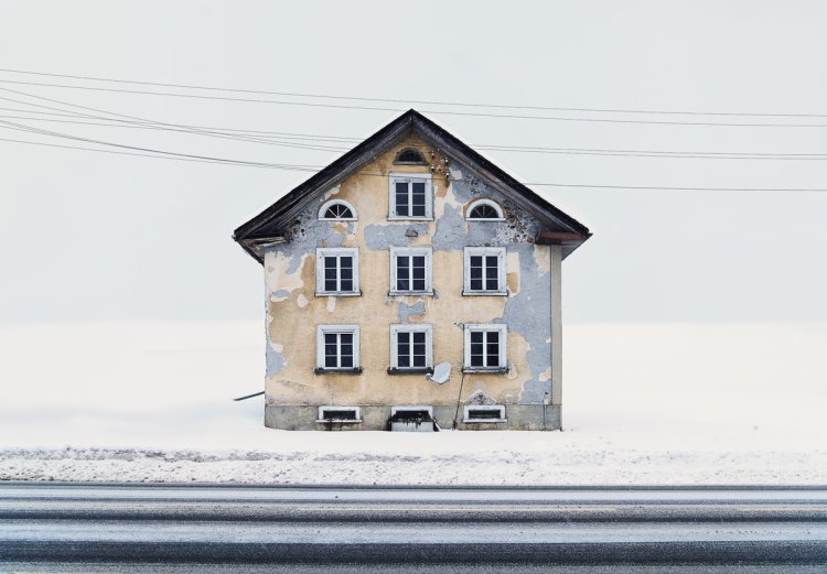 Kleines Schweizer Haus mit abbröckelndem, gelbem Putz an der Fassade, darunter kommen graue Flecken oder sogar das Steingemäuer zum Vorschein. Der Hintergrund ist weiss und verschneit, im Vordergrund eine angeschnittene Strasse und Hochspannungsleitungen.