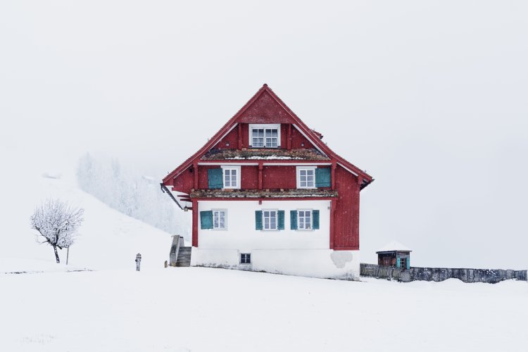 Ein kleines Haus mit weisser Fassade unten und roter Fassade unter dem Dach. Rechts führt eine Mauer aus dem Bild, die Landschaft rundherum ist verschneit und verschwindet im Nebel.