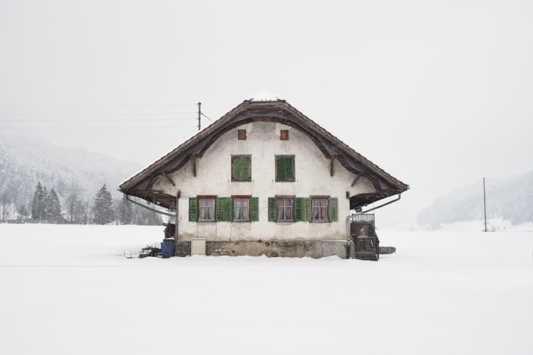 Kleines Schweizer Haus mit rund geschwungenem Dach, weisser Fassade mit Sprüngen und grünen Fensterläden. Dahinter ein nebeliger, verschneiter Hintergrund.