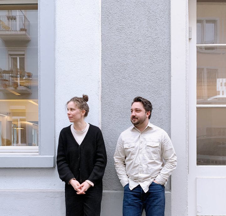 Lisa Mäder in schwarzem Pullover und Hubert Holewik in beigem Hemd stehen vor einer Hausmauer und schauen nach links