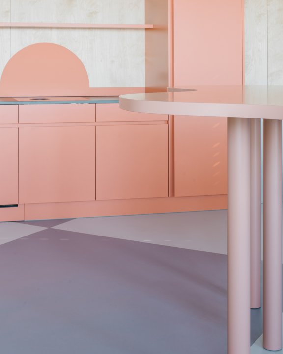 Korallefarbige Küche im Hintergrund, vorne rechts ein angeschnittener rosa Stehtisch auf einem violetten Boden
