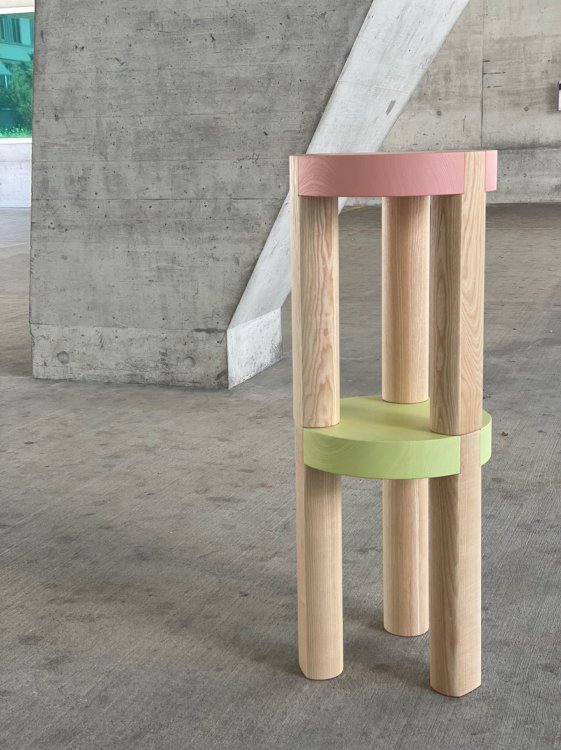 Zwei einfache Holzhocker mit einer rosa und einer grünen Sitzfläche stehen aufeinander, im Hintergrund Beton
