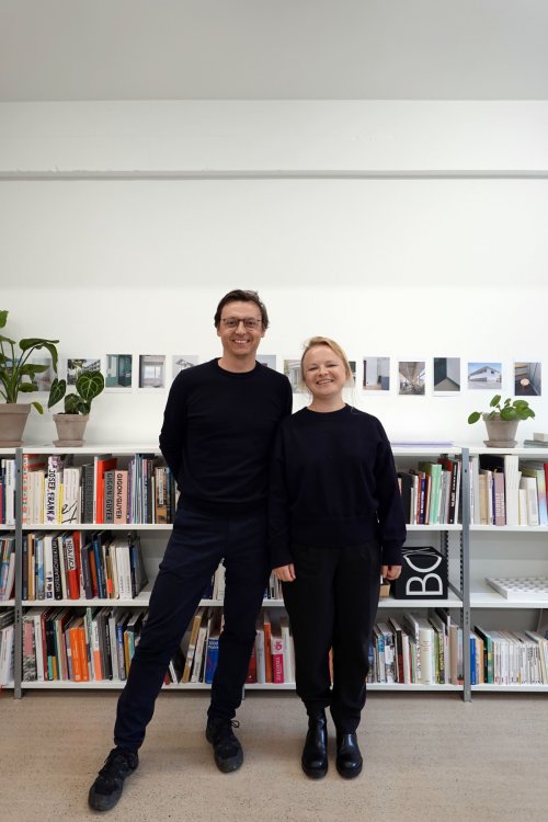 Porträt eines Architekt:innenpaars, beide in Schwarz, vor einem Bücherregal