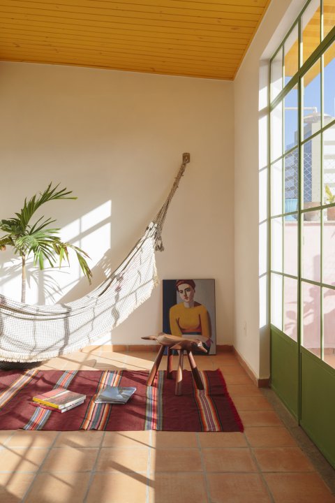 Wohnraum mit grossflächigem, grün gerahmten Fenster rechts, einer Hängematte über weinrotem Teppich rechts und gelber Decke.