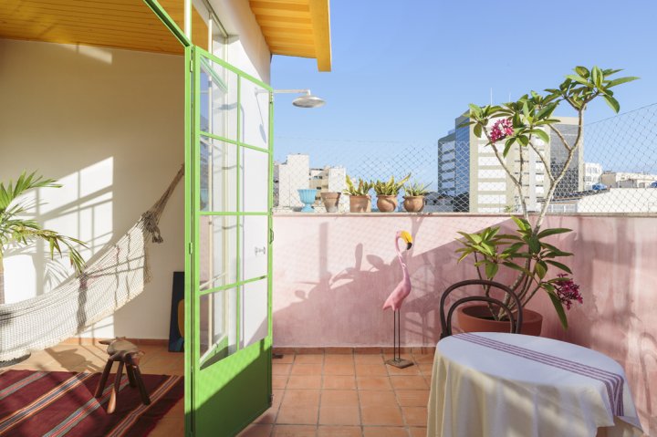 Balkon mit pink gestrichenen Wänden, dahinter Stadt mit blauem Himmel, links angeschnitten Einblick in ein Wohnzimmer.
