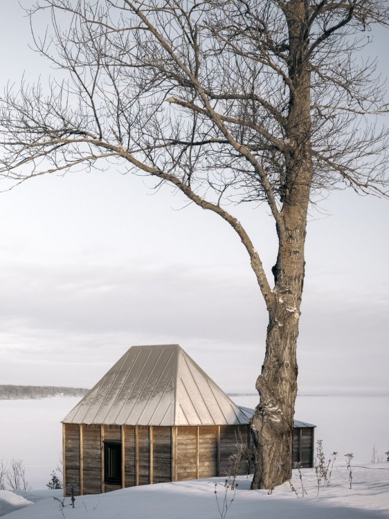 Ein kleines Holzhaus steht neben einem blätterlosen Baum an einem See in einer verschneiten Winterlandschaft.