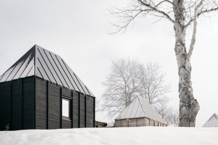 Perspektive von unten auf einen Hügel, wo ein kleines, schwarzes, holzverkleidetes Haus steht, im Hintergrund angeschnitten weitere Häuser der gleichen Typologie, vorne rechts im Bild ein Baum, alles in einer vernebelten Schneelandschaft.