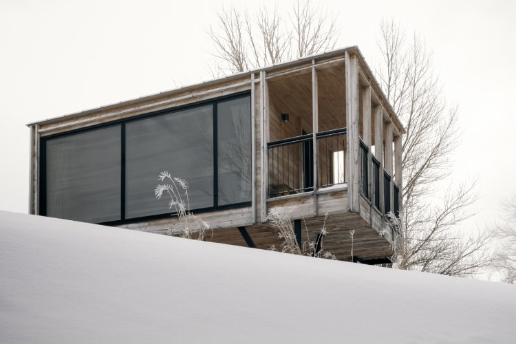 Ein hölzernes Tiny House mit grosser, frontaler Fensterscheibe steht oben auf einem verschneiten Hang.