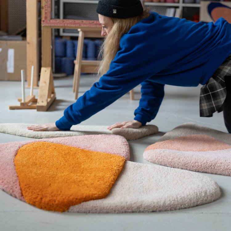 Eine Frau mit Mütze und blauem Pullover kniet auf dem Boden mit den Händen auf mehreren bunten Teppichen.