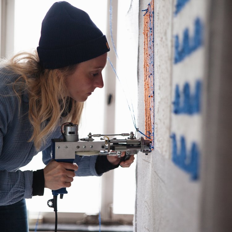 Eine Frau mit Mütze und blonden Haaren arbeitet mit einer Tufting Maschine an einem Wandteppich, der vor ihr hängt.