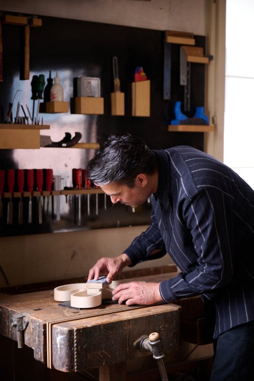 Der Künstler Christian Neuenschwander bei der Arbeit in seinem Atelier. Er trägt ein dunkel gestreiftes Hemd und konzentriert sich darauf, eine helle, geschwungene Holzskulptur an einer Werkbank von Hand zu schleifen. Im Hintergrund ist eine Wand mit verschiedenen Werkzeugen wie Stechbeiteln und Zwingen zu sehen.