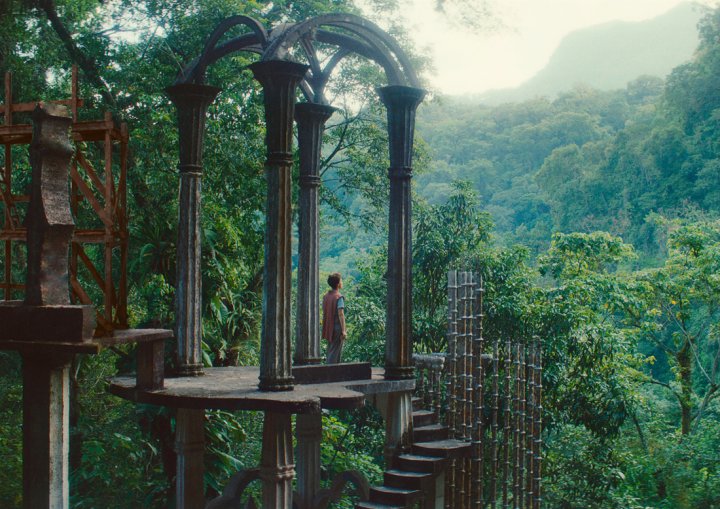 Filmstill aus Leonora in the Morning Light: Eine Person steht auf einer surrealen Betonplattform mit Bögen und Treppen in Las Pozas, umgeben von dichtem, grünem Regenwald und Bergen im Hintergrund.