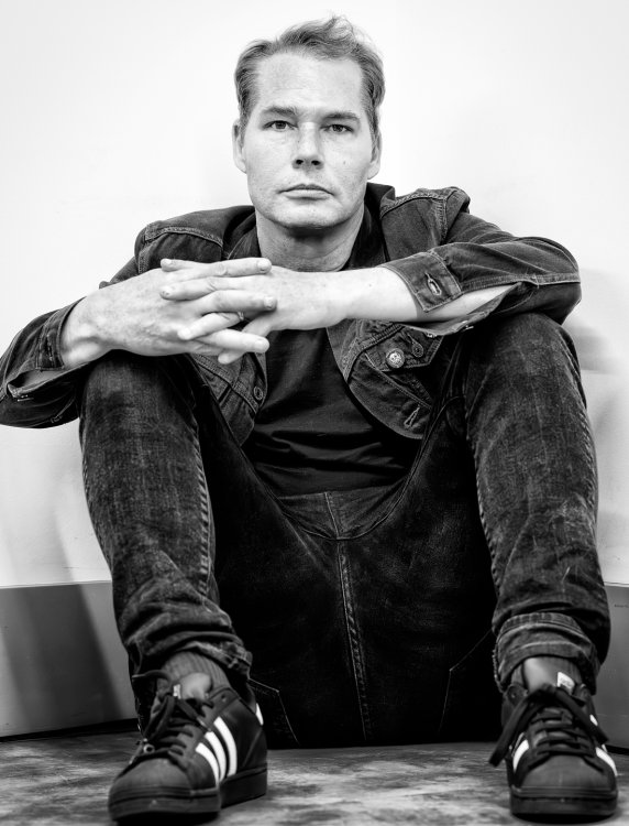 Schwarzweisses Porträt von Shepard Fairey, sitzend mit verschränkten Händen, in Jeansjacke, Jeans und Turnschuhen, frontal in die Kamera blickend.