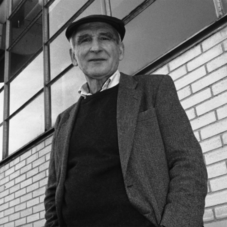 Schwarz-Weiss-Porträt des Designers Kurt Thut vor einer modernen Glasfassade und einer Backsteinwand; er trägt eine Schiebermütze, Sakko und Pullover.