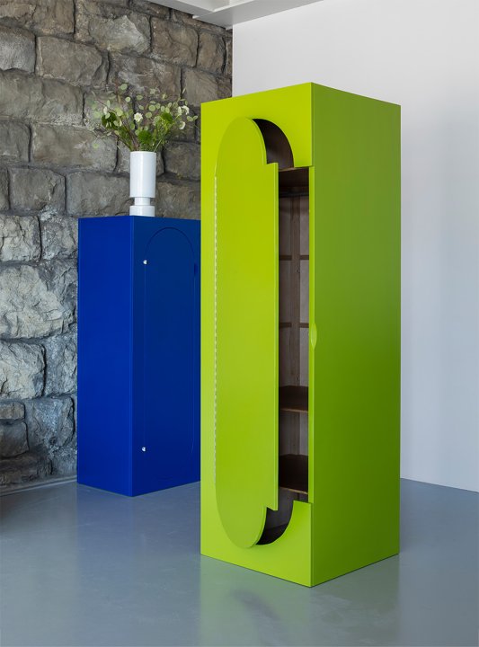 Schrank „Squadra“ von Klaus Vogt: hoher, limegrüner Schrank mit geöffneter Tür und sichtbaren Regalböden; im Hintergrund ein blauer Schrank vor einer Steinwand.