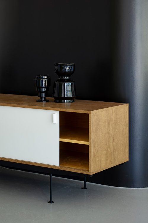 Detailansicht des Sideboards „Sesam“ von Dieter Waeckerlin: Eichenholz-Korpus mit einer weissen Schiebetür und offenen Regalfächern, dekoriert mit schwarzen Vasen vor einer dunklen Wand.