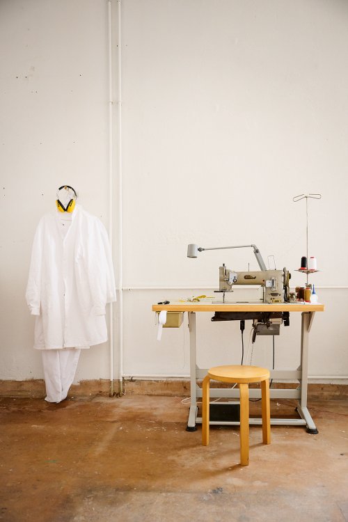 Blick in ein helles Atelier mit einer industriellen Nähmaschine auf einem Arbeitstisch und einem davorstehenden Holzhocker. An der weissen Wand links hängt ein weisser Arbeitskittel zusammen mit einem gelben Gehörschutz. Die Szene wirkt minimalistisch und funktional vor einem rustikalen Werkstattboden.