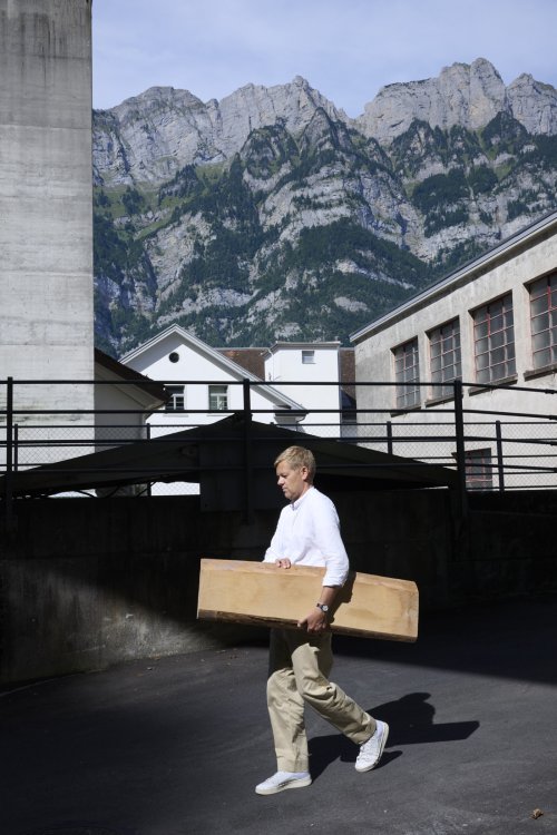 Der Designer Ville Kokkonen trägt ein grosses, massives Holzbrett über einen asphaltierten Aussenbereich vor seinem Atelier. Im Hintergrund ragen markante, felsige Berggipfel unter einem blauen Himmel empor, während die Architektur der umliegenden Gebäude den industriellen Charakter des Standorts betont.