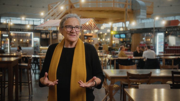 Porträt der Architektin Barbara Buser in der Basler Markthalle. Sie trägt ein schwarzes Oberteil, eine Brille und einen markanten gelben Schal, während sie mit erklärenden Handgesten spricht. Im Hintergrund ist die belebte Halle mit Holztischen, Marktständen und Gästen in einer warmen, leicht unscharfen Atmosphäre zu sehen.