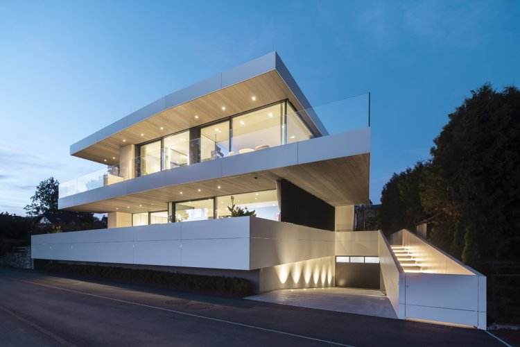 Moderne, mehrstöckige Villa am Attersee bei Dämmerung mit beleuchteter Fassade aus Dekton-Paneelen. Die Architektur besticht durch weit auskragende Terrassen mit Glasgeländern, eine integrierte Tiefgarage und eine elegant beleuchtete Aussentreppe, die das luxuriöse Design bei Nacht betonen.