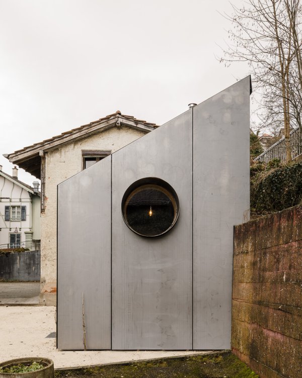 Rückansicht der asymmetrischen Giebelseite des Tiny Houses in Münchenstein. Die Fassade ist bündig mit vertikalen, matten Stahlpaneelen verkleidet. Im Zentrum befindet sich ein kreisrundes Bullauge-Fenster, durch das im Inneren eine brennende Glühbirne sichtbar ist. Rechts begrenzt eine rötliche Betonmauer das Grundstück.