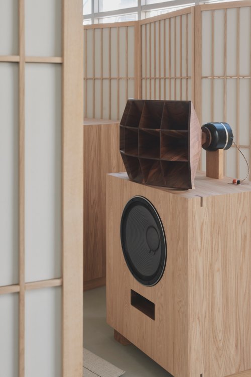 Detailaufnahme eines grossformatigen Lautsprechers aus hellem Massivholz mit einem aufgesetzten, komplex gefertigten Horn-Vorsatz aus dunklerem Holz. Die Konstruktion steht vor einem Hintergrund aus japanischen Paravent-Raumteilern und verdeutlicht die präzise Holzverarbeitung von Karimoku Furniture.
