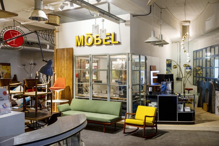 Blick in das «H100 Openstorage» in Zürich: Ein weitläufiges, industriell anmutendes Möbellager mit einer kuratierten Auswahl an Designklassikern. Im Vordergrund stehen ein grünes Sofa und ein gelber Sessel, während im Hintergrund ein gläserner Büroraum unter einem grossen gelben «MÖBEL»-Logo zu sehen ist. Diverse Leuchten und Vintage-Schilder unterstreichen den Charakter als Inspirationsquelle.
