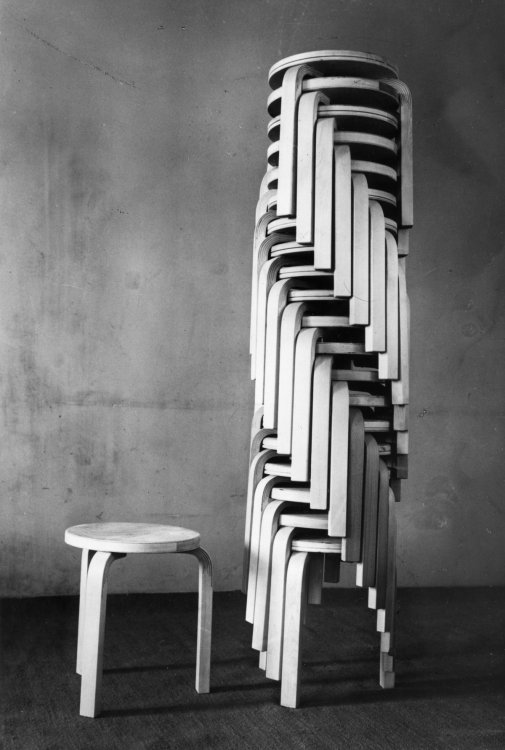 Historische Schwarz-Weiss-Fotografie eines hohen Stapels von Artek Hocker 60 vor einer kargen Wand. Ein einzelner Hocker steht separat daneben. Die Aufnahme betont die Stapelbarkeit und die ikonischen L-Beine aus gebogenem Holz, die spiralförmig ineinandergreifen.