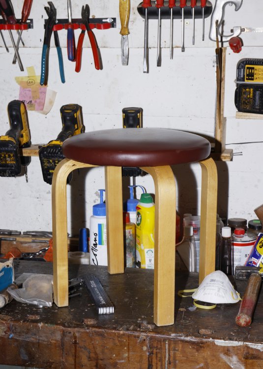 Ein klassischer Artek Hocker mit dunkler Lederpolsterung und den typischen L-Beinen aus hellem Holz steht auf einer rustikalen Werkbank. Im Hintergrund ist eine Werkstattwand mit Handwerkzeugen wie Zangen, Schraubenziehern, Bohrmaschinen und Reinigungsmitteln zu sehen.