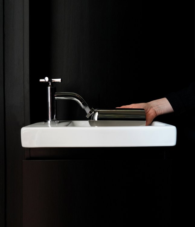 Eine atmosphärische Nahaufnahme zeigt, wie das glänzende Metallgefäss „Tilt“ von Altherr Weiss unter dem flachen Auslauf einer modernen Chrom-Armatur in einem weissen Waschbecken befüllt wird. Eine Hand hält das Objekt waagerecht, um die funktionale Handhabung in einem dunklen, eleganten Interieur mit schwarzer Wand zu demonstrieren.