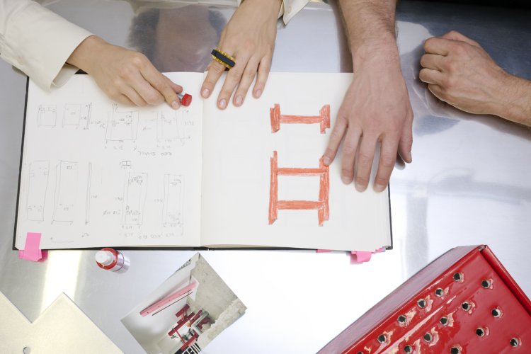 Draufsicht auf einen Arbeitstisch aus Edelstahl, an dem das Designduo Altherr Weiss gemeinsam an einem Skizzenbuch arbeitet. Man sieht ihre Hände über den Seiten, die technische Zeichnungen und eine markante rote Skizze einer leiterähnlichen Struktur zeigen; auf dem Tisch liegen zudem ein Entwurfsfoto, Farbmuster und kleine Werkzeuge, die den kreativen Designprozess dokumentieren.