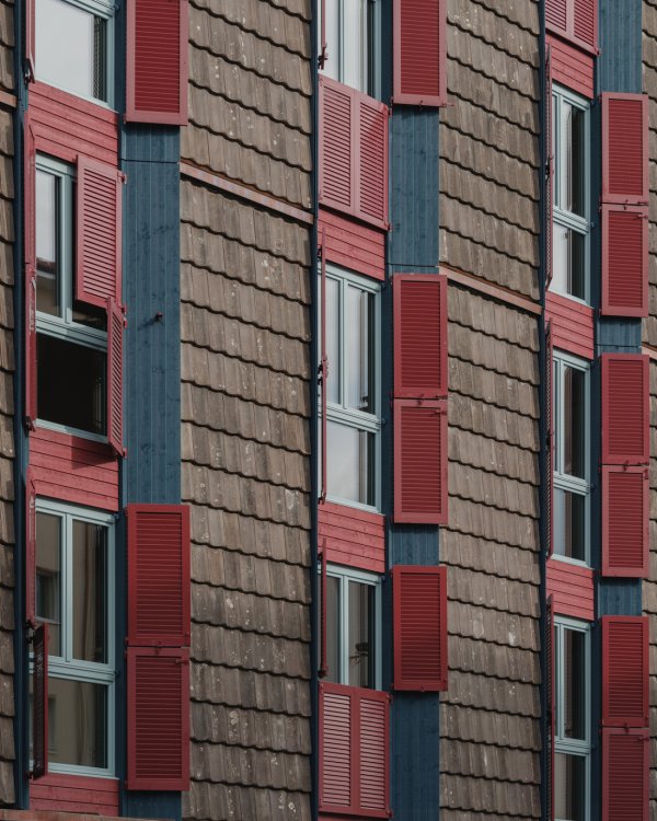 Detailaufnahme der Fassade des Wohnhauses LysP8. Sie zeigt die markante Kombination aus vertikal angeordneten, gebrauchten braunen Dachziegeln und dunklen Holzelementen, ergänzt durch Fenster mit auffälligen roten Klappläden.