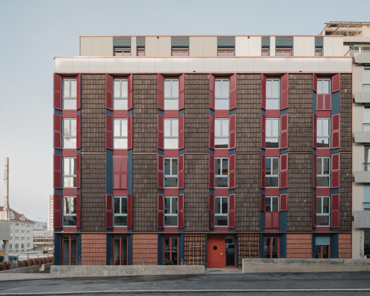Vorderansicht des Wohnhauses LYSP8 von Loeliger Strub Architektur mit einer strukturierten Rasterfassade. Die Aussenwände bestehen aus wiederverwendeten braunen Dachziegeln und blauen Holzelementen, ergänzt durch hohe Fenster mit auffälligen roten Klappläden.