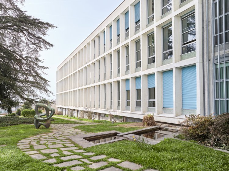 Seitenansicht der hellen Rasterfassade des «Le Cèdre» in Lausanne. Im gepflegten Aussenbereich sind ein gepflasterter Weg, zwei rechteckige Wasserbecken und eine bronzene Skulptur zu sehen. Die grossen Fenster der Büroetagen sind teilweise durch blaue Lamellenstoren verschlossen, was einen farblichen Akzent zur weissen Betonstruktur setzt.
