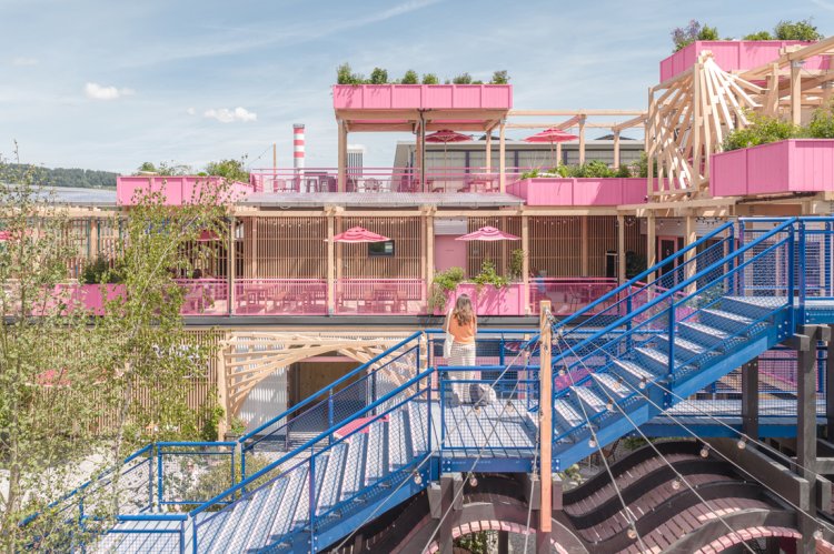 Luftaufnahme der Remise Rosa in Zürich West bei Dämmerung: Eine vielschichtige, mehrgeschossige Holzkonstruktion mit rosa Akzenten, Dachterrassen, Pflanzen und Gästen inmitten einer urbanen Umgebung mit Kränen und modernen Gebäuden.